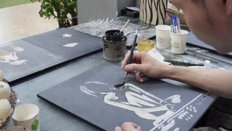 Nghệ nhân đang tiến hành vẽ phác thảo hoặc tô màu cho chi tiết trên bề mặt tranh sơn mài khảm trai